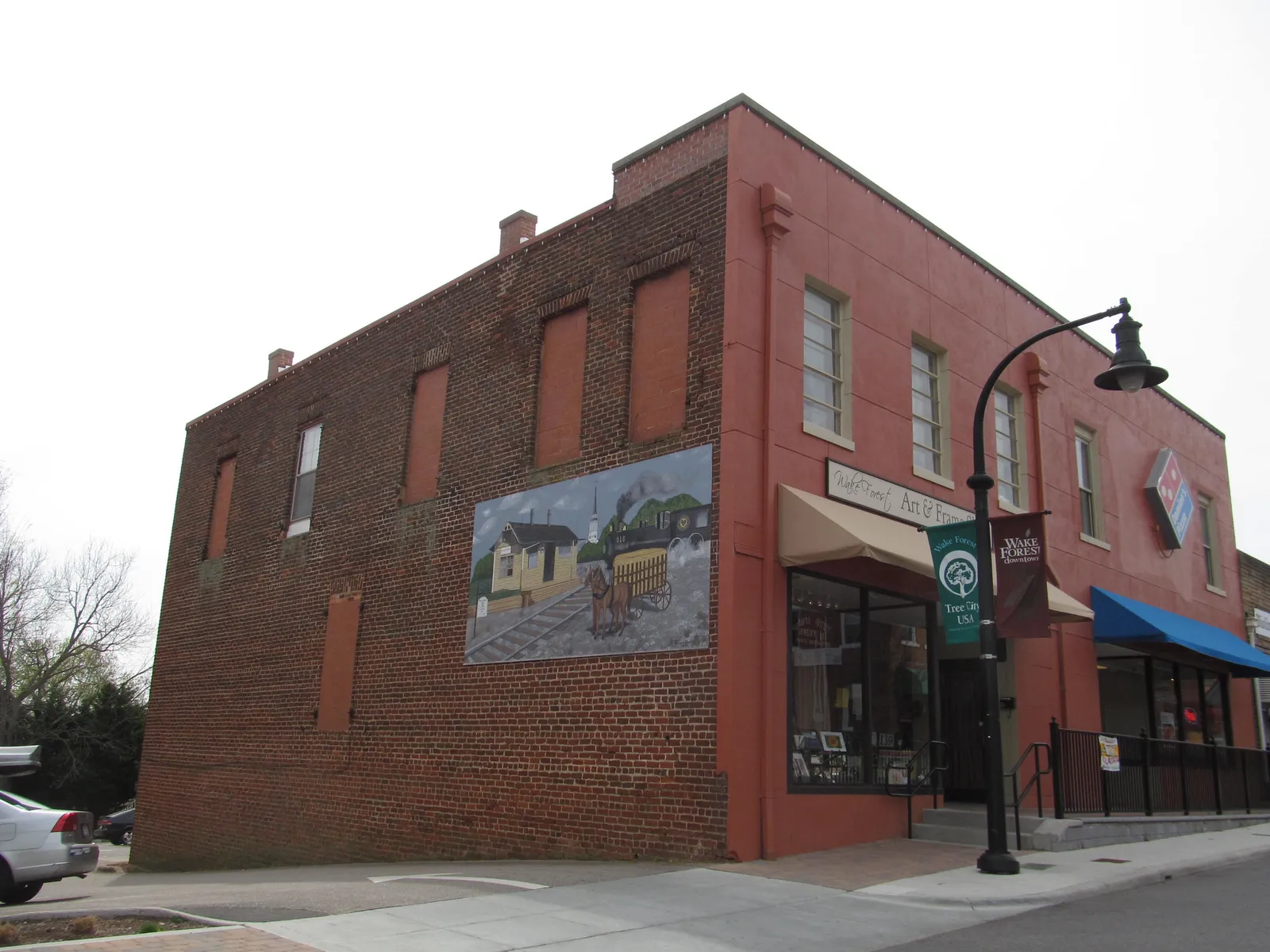 Storefronts in a brick building in downtown Wake Forest, with a painted mural of a train and depot on its side wall.